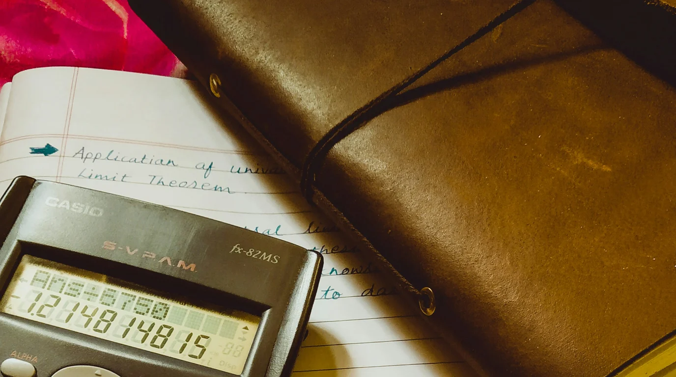 A scientific calculator showing a long result, resting on a lined notebook of handwritten notes beside a leather-bound journal.