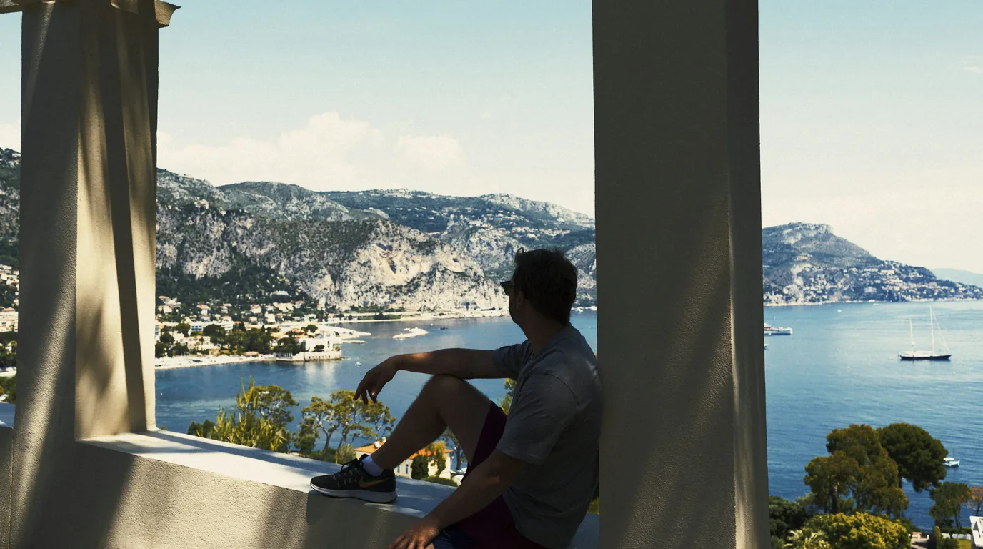 A man sitting on a terrace wall between two pillars, looking out over a Mediterranean bay with yachts and a headland beyond.