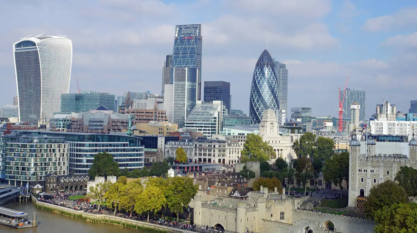 Glass and steel towers standing behind a riverside stone castle and leafy gardens, seen from above under a pale blue sky.